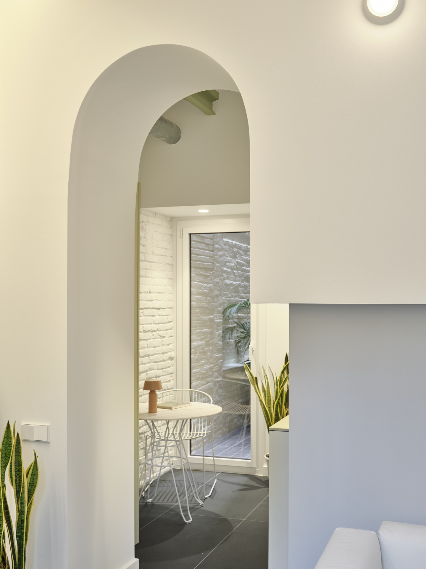 Arched opening in CRÜ’s “La Dolors” renovation framing a small dining nook with wire chairs, a round table, and a view toward the whitewashed brick patio.